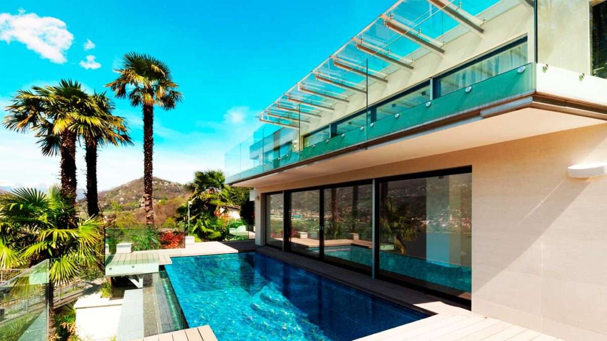 Banner mostrando uma casa de manhã, no verão com uma piscina de vidro, portas de vidro separando a piscina do ambiente interno e no andar de cima um guarda corpo de vidro.