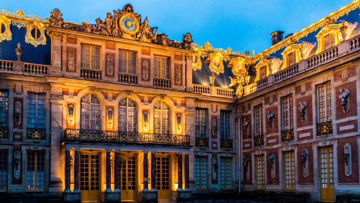 Banner mostrando a parte da frente do Palácio de Verseilles e seus vidros. Visto a noite com a iluminação no pé da edificação.